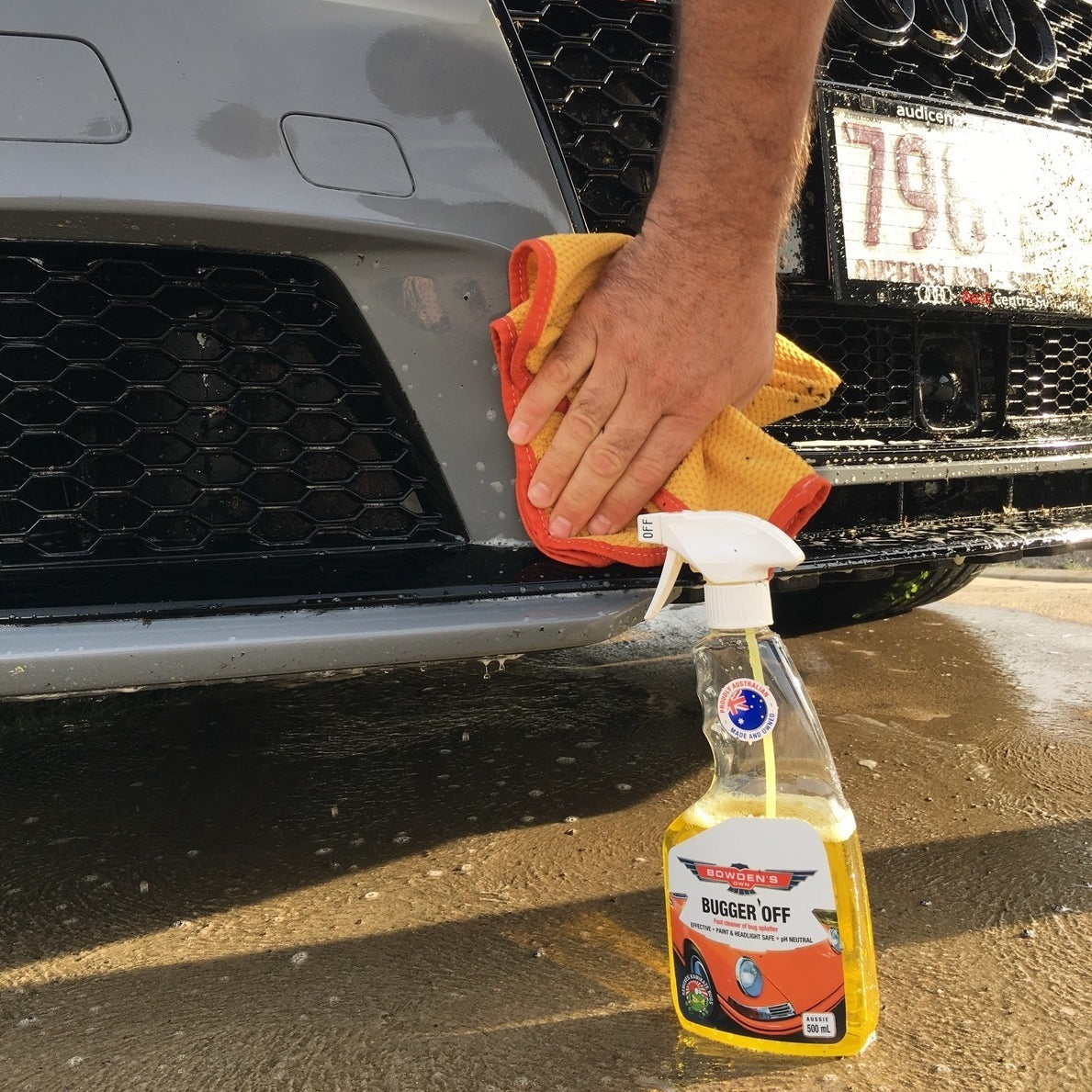 Person cleaning a car with a spray bottle labeled 'Buggers Off' on a sunny day.