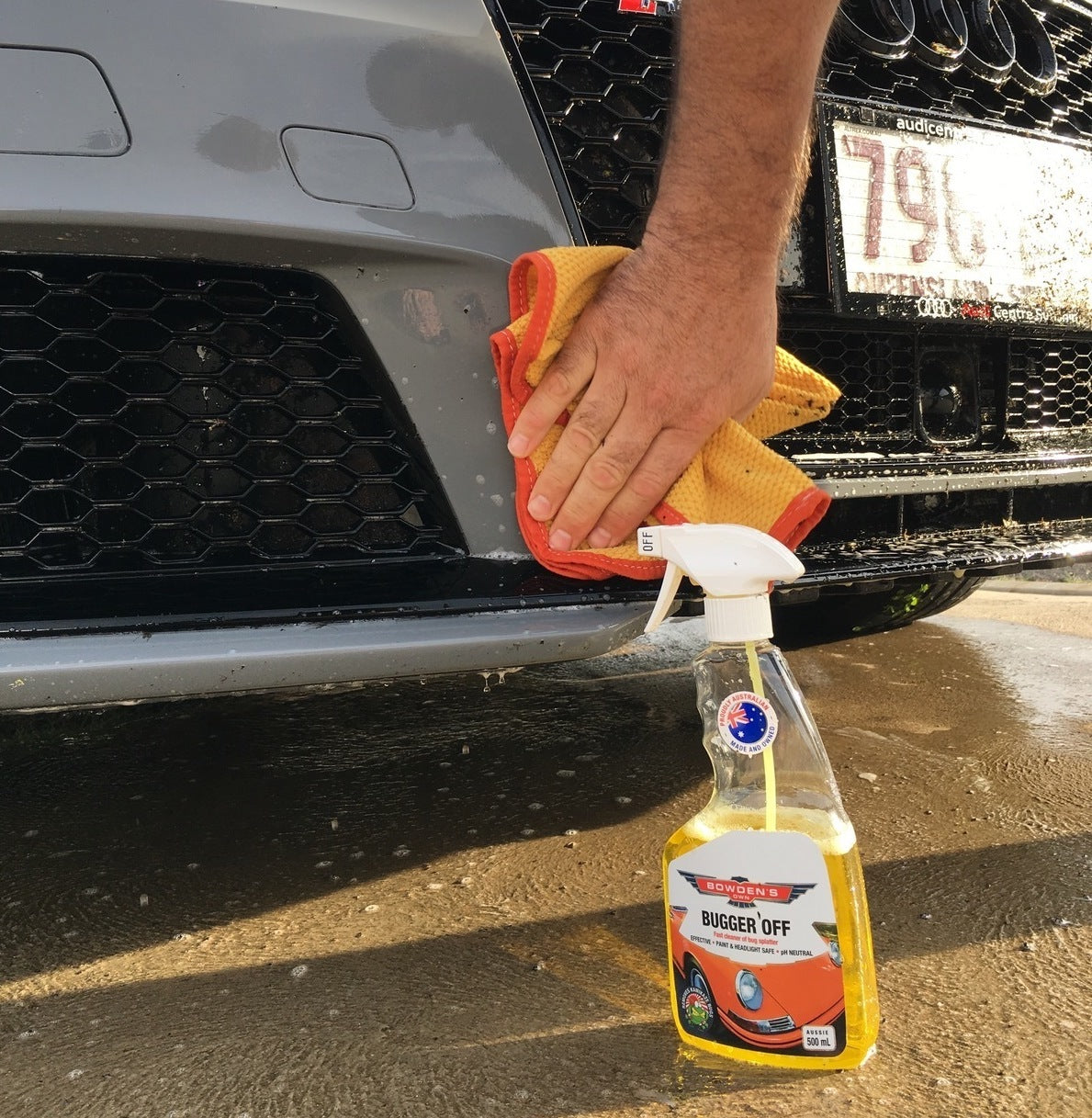 Person cleaning a car with a spray bottle labeled 'Buggers Off' on a sunny day.
